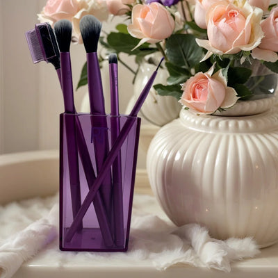 Real Techniques makeup brushes in a purple holder, surrounded by pink roses, on a plush surface.