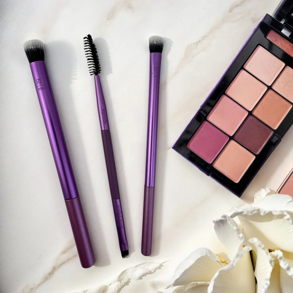 Real Techniques makeup brushes set on a marble surface with an eyeshadow palette, featuring purple handles and diverse brush shapes.