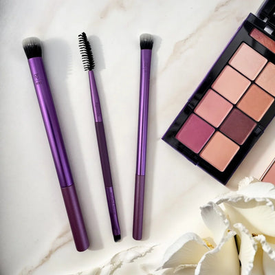 Real Techniques makeup brushes set on a marble surface with an eyeshadow palette, featuring purple handles and diverse brush shapes.