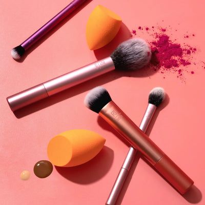 Real Techniques makeup brush set with blending sponges and powder, showcasing vibrant colors and textures on a coral background.